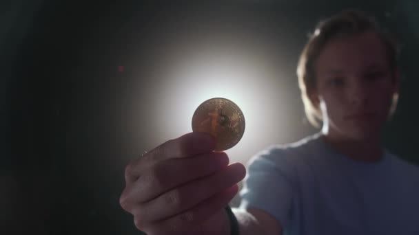 Un joven sostiene un bitcoin en su mano, a través del cual la luz brilla a través de. — Vídeo de stock
