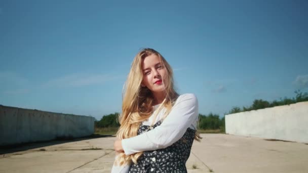 Ein junges Mädchen in einem schönen Kleid steht in der Nähe von Betonmauern und einem Feld. Ein starker Wind bläst in die Haare. — Stockvideo