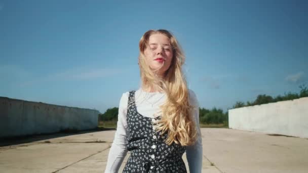 Ein junges Mädchen in einem schönen Kleid steht in der Nähe von Betonmauern und einem Feld. Ein starker Wind bläst in die Haare. — Stockvideo