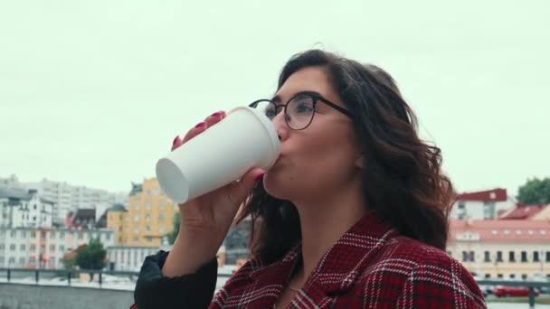 En ung flicka dricker kaffe och håller sig till sin telefon. Flickan löser viktiga saker i den friska luften med en kopp kaffe i händerna. — Stockvideo