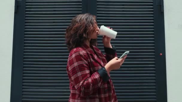 Une jeune fille boit du café et colle à son téléphone. La fille résout des choses importantes dans l'air frais avec une tasse de café dans ses mains. — Video