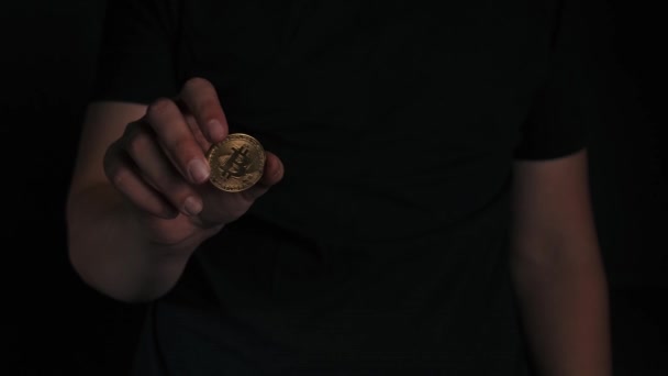 Mans χέρι κατέχει και λάμπει χρυσό Bitcoin σε μαύρο φόντο. νέο μεταλλικό κρυπτονόμισμα στα δάχτυλα. πρόσωπο δείχνει το μελλοντικό νόμισμα. online εξόρυξη και εμπορία — Αρχείο Βίντεο