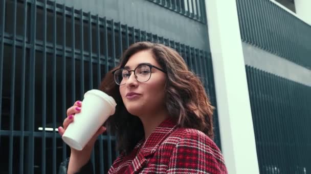 Una joven bebe café al aire libre. Una chica feliz está tomando café cerca del centro de negocios. — Vídeos de Stock