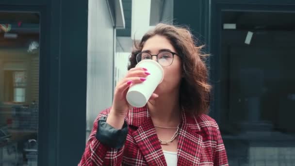 Ein junges Mädchen trinkt Kaffee an der frischen Luft. Ein glückliches Mädchen trinkt Kaffee in der Nähe des Geschäftszentrums. — Stockvideo