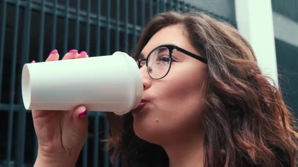 En ung flicka dricker kaffe i friska luften. En lycklig flicka dricker kaffe nära affärscentret.. — Stockvideo