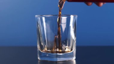 Orta çerçeve, mavi arkaplanda bardağa dökülen bir kola kutusunu tutan bir elin yavaş çekim görüntüsüdür..