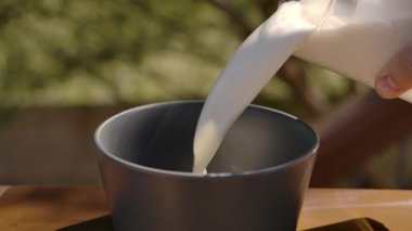 Güneşli bir günde, yaz mutfağındaki bir kaseye dökülen taze sütün yavaş çekimde yakın plan çekimi..