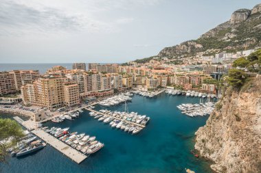 Lüks yatlar, turkuaz Akdeniz suları, modern liman mimarisi ve bulutlu bir gökyüzü altındaki tepe kenti manzarasının geniş panoramik manzarası