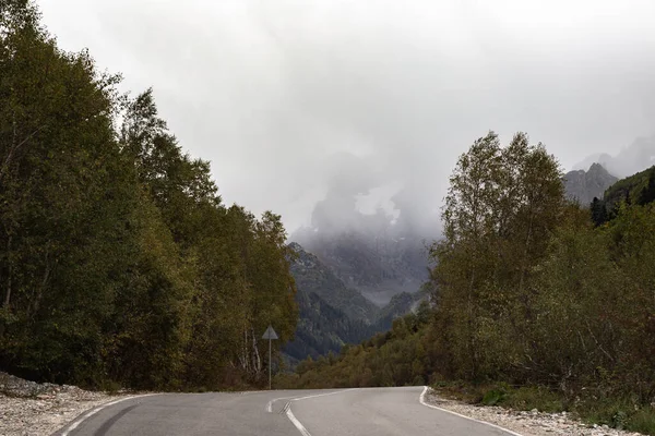 Gama de montanhas cobertas de neve nas nuvens. Estrada. Floresta densa nas encostas das montanhas Cáucaso, Rússia — Fotografia de Stock