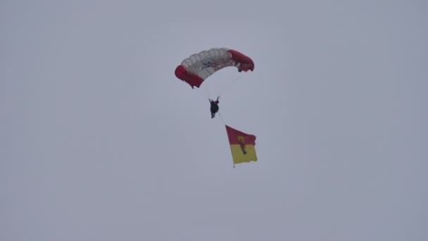 Paraquedistas com paraquedas vermelho e branco e uma bandeira pousando em um céu cinzento nublado — Vídeo de Stock