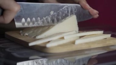 Mutfakta, duvarları mora boyanmış, tahta bir tahtanın üzerinde, çok büyük, keskin bir bıçakla peynir kesen bir kadın eli. Yandan çekilen Yemek