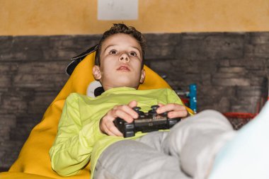 Çocuk, elinde kumandası olan sarı bir tişört giyerek bir puf üzerinde video oyunları oynuyor. Yaşam biçimi