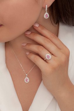 Beyaz elbiseli kahverengi saçlı bir kadın çıplak tırnak cilası eliyle yüzük ve küpe şeklinde bir elmas kolye gösteriyor. İnternette satılık.