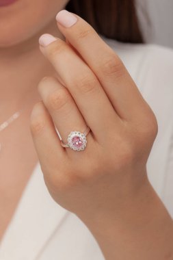 Beyaz elbiseli ve kumral saçlı kadın model elinde pembe bir parçayla halka şeklinde elmas bir yüzükle poz veriyor..