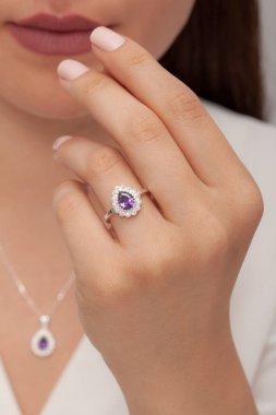 Beyaz tenli, bakımlı, beyaz elbiseli kadın, mor bir kolye ve yüzük sergiliyor..