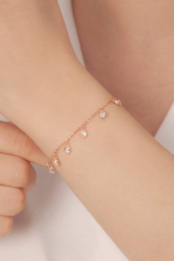 Beyaz elbiseli beyaz tenli bir kadın beyaz taşlı gül renkli bir bileklikle poz veriyor. İnternette satılık.