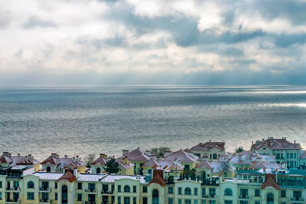 Bulutlu havada denizin arka planına bakan evler ve çatılar, denizin üzerindeki dramatik kara bulutlar boyunca güneş ışınları yol alır..