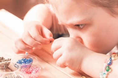 Küçük bir kız dikiş işiyle meşgul, kendi elleriyle mücevher yapıyor, ipliğe çok renkli boncuklar diziyor..