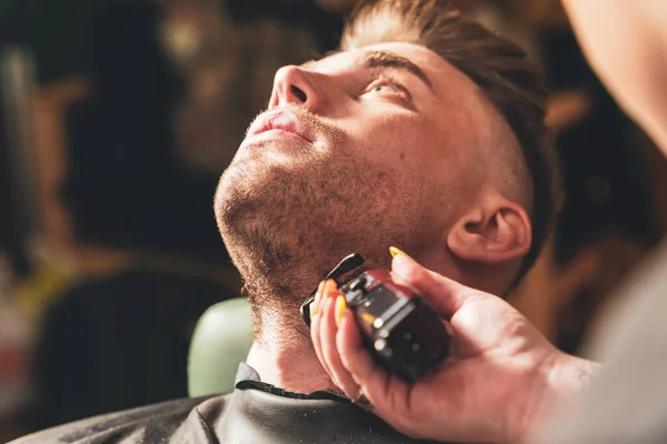 Bearded man in a barbershop, hands of a hairdresser with a beard clipper, close up.