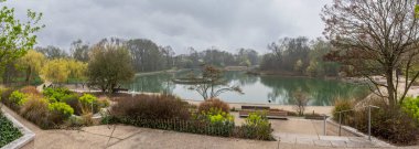 Gennevilliers, Fransa - 03 08 2026: Chanteraines Parkı. Bahar mevsiminde doğa. Ağaçların ve bitki örtüsünün panoramik görüntüsü bulutlu gökyüzü ile göle yansıyor