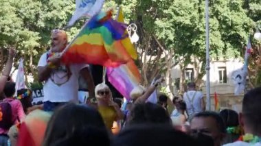 Güneş gözlüklü mutlu adam kalabalığın üzerinde gökkuşağı bayrağı sallıyor.