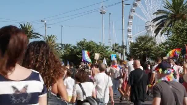 Kalabalık, bir dönme dolaba yakın caddede yürürken gökkuşağı bayrakları sallayarak kentte ilerliyor. — Stok video