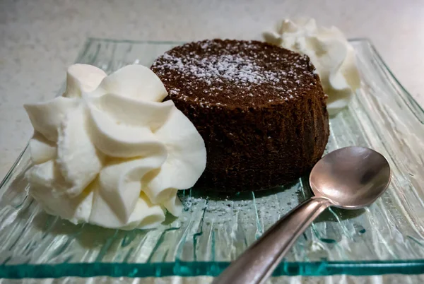 Üzerinde pudra şekeri ve yanında krema olan küçük çikolatalı pasta, cam tabakta servis edilir.