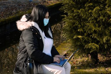 Cerrahi maskeli bir kadın parkta dizüstü bilgisayarla çalışıyor. Koronavirüs nedeniyle açık havada telsiz çalışıyor. Çevrim içi iş.
