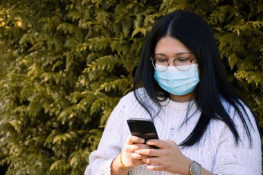 Sosyal ağlarda cep telefonunu kullanan cerrahi maskeli bir kadın. Parktaki kadın..