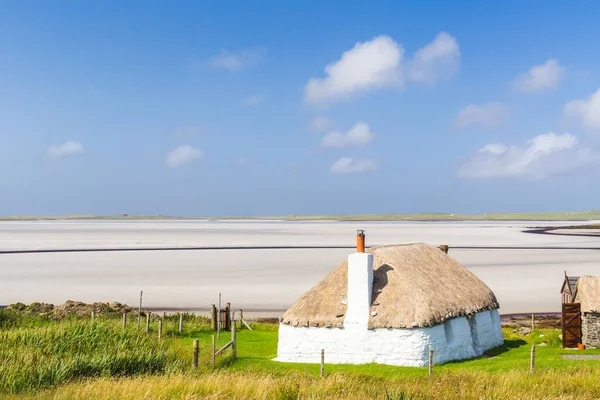 Chalet blanc traditionnellement construit avec toit de chaume, à côté de la baie turquoise, avec un ciel sombre nuageux orageux au-dessus. Île de North Uist, Écosse — Photo