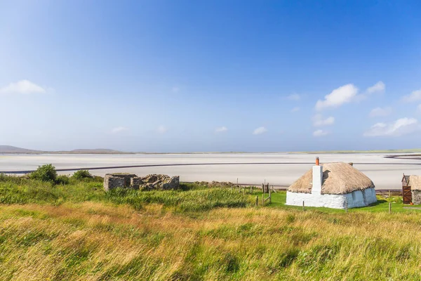 Traditionellt byggd vit stuga med halmtak, intill turkosa viken, med stormiga molniga mörka himmel ovan.ön North Uist, Skottland — Stockfoto