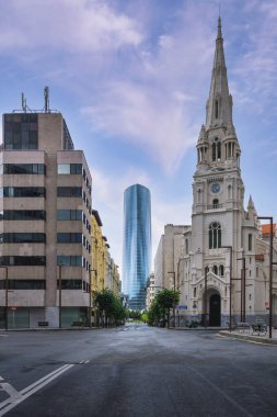 İspanya 'nın Bilbao kentindeki sakin bir şehir caddesi modern cam gökdelenle parçalı bulutlu gökyüzünün altındaki tarihi bir kilise arasındaki zıtlığı gösteriyor..