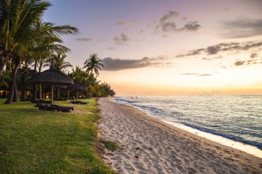 Hint Okyanusu, Mauritius sahilinde palmiye ağaçları, sazdan şemsiyeler ve yumuşak günbatımı ışıklarıyla tropik bir sahil.