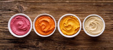 Ahşap arka planda pancar, balkabağı ve kırmızı biber gibi çeşitli sebzeler içeren farklı renkte humus seti.