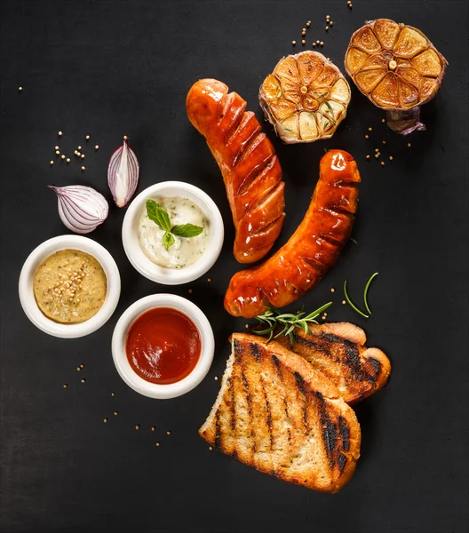 Salchichas a la parrilla con diferentes tipos de salsas sobre un fondo negro —  Fotos de Stock