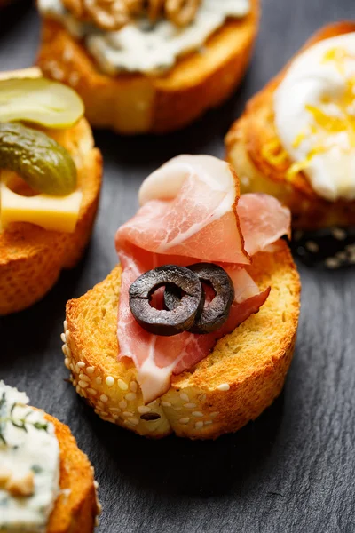 Canapé pequeño con baguette a la parrilla con la adición de jamón, aceitunas sobre fondo negro — Foto de Stock