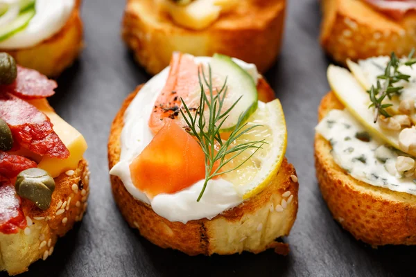 Small canape with grilled baguette with the addition of cremy cheese, smoked salmon, cucumber, lemon and dill on black background — Stock Photo, Image