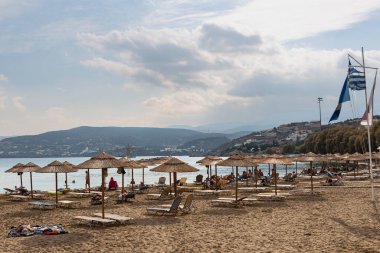 Kumlu sahil, güneşli şezlonglar, kamış şemsiyeleri ve tatilciler denizin, dağların ve mavi gökyüzünün önünde. Yunanistan. Agios Nikolaos