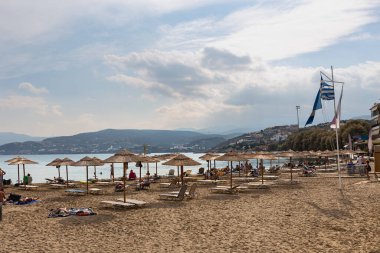 Kumlu sahil, güneşli şezlonglar, kamış şemsiyeleri ve tatilciler denizin, dağların ve mavi gökyüzünün önünde. Yunanistan. Agios Nikolaos