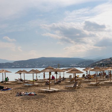 Kumlu sahil, güneşli şezlonglar, kamış şemsiyeleri ve tatilciler denizin, dağların ve mavi gökyüzünün önünde. Yunanistan. Agios Nikolaos