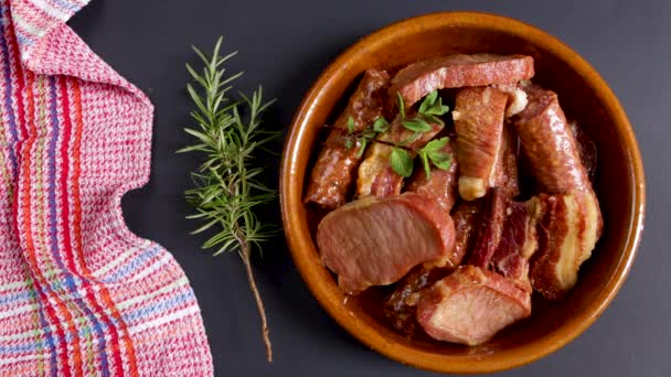黒い背景にローズマリーの小枝と粘土鍋に保存された豚肉の肋骨 ロース肉と豚肉のソーセージ 典型的なスペイン料理 — ストック動画