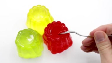Çeşit çeşit kırmızı, yeşil ve sarı jöleler beyaz, çilekli, limonlu, limonlu, jöleli, kaşıklı bir el bir porsiyon alır.