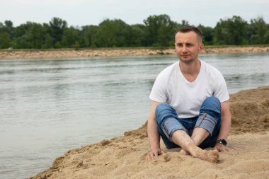 Sahildeki nehirde oturan yalnız, pozitif ve gülümseyen adam.