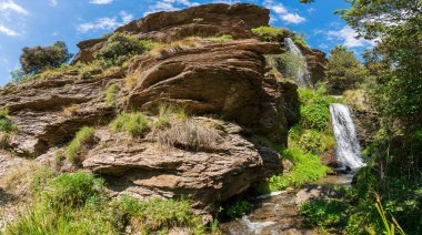Sierra Nevada 'da bir vadide şelale, çimenler ve ağaçlar var, kayalar var ve gökyüzünde bulutlar var.