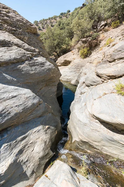 Water flowing down a ravine in Sierra Nevada, there is a pool of crystal clear water, it is a rocky area, there is grass on the rock, the sky is clear