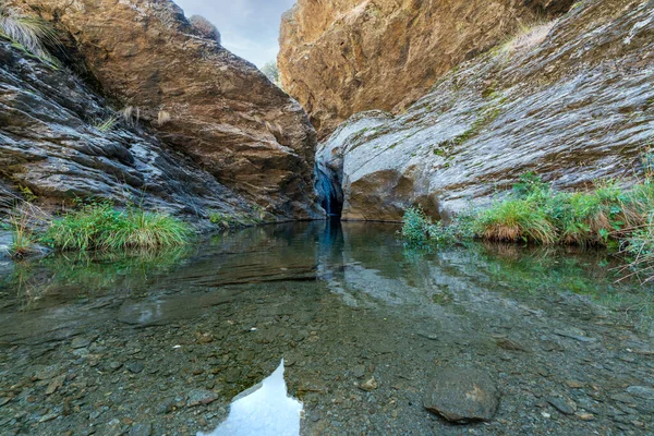 Water flowing down a ravine in Sierra Nevada, there is a pool of crystal clear water, it is a rocky area, there is grass on the rock, the sky is clear