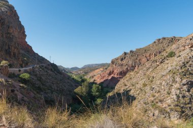 Güney İspanya 'nın dağlık arazisi, dağların çalıları, dağ yamacı boyunca uzanan bir yol, yüksek voltajlı kuleler, kavak ve kamış ormanları var, gökyüzü açık.