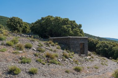 Taştan yapılmış küçük bir ev, ağaçlar ve çalılar var, taşlar var ve gökyüzü temiz.