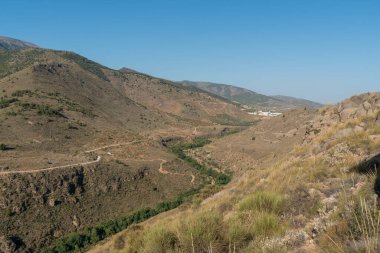 Güney İspanya 'nın dağlık arazisi, ağaçlar ve çalılar var, alan engebeli, uzakta bir kasaba var, gökyüzü açık.