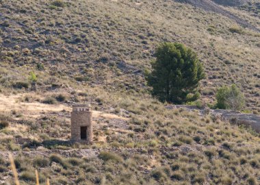 Bir dağ yamacında bir inşaat ve birkaç ağaç, yapı taştan yapılmış, yerde taşlar var, çalılar var.